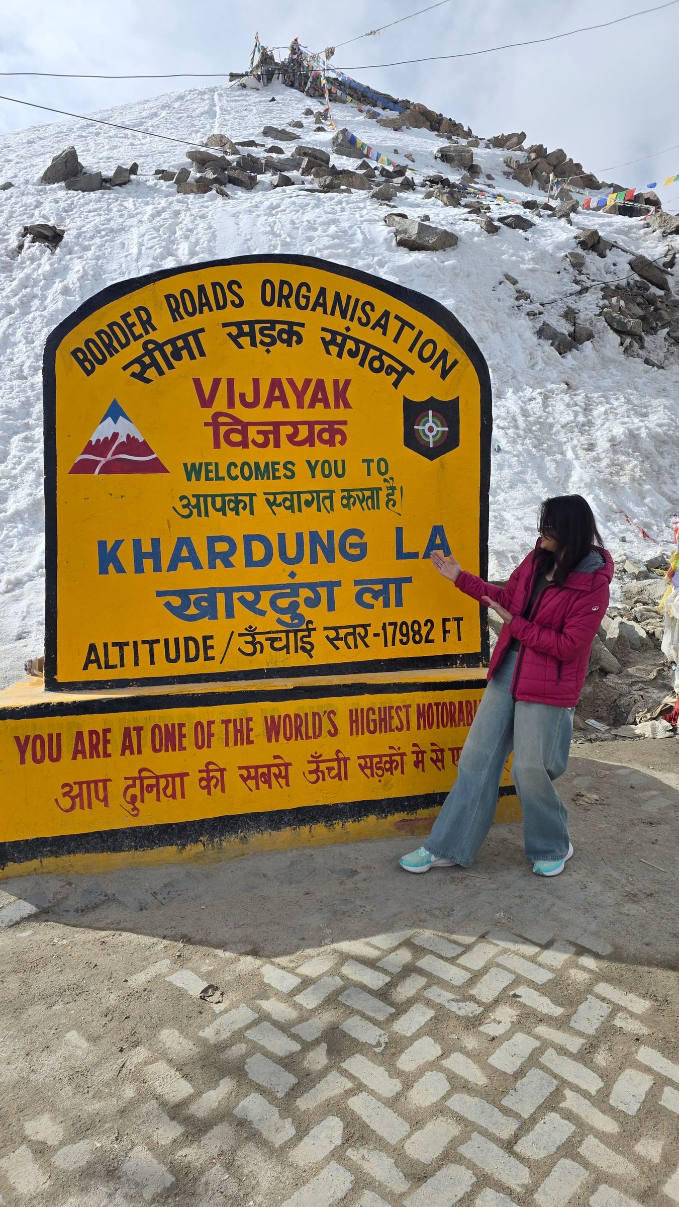A traveller gesturing toward the yellow Khardung La summit sign at the pass